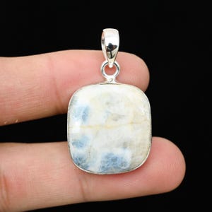 May include: A square pendant with a silver bail. The pendant features a cream-colored stone with blue and yellow inclusions. The stone is set in a silver bezel. The background is black.