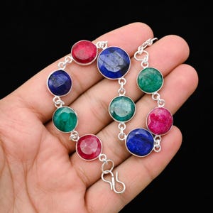 May include: A silver bracelet featuring a series of round, faceted gemstones in shades of red, blue, and green. The gemstones are set in individual silver bezels and connected by small silver links. The bracelet is displayed against a black background.