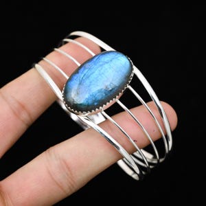 Peut inclure: Un bracelet manchette en argent orné d'une grande pierre ovale de labradorite aux reflets bleus et irisés. La pierre est sertie dans une lunette en argent et entourée de barres d'argent parallèles. Le bracelet est présenté sur fond noir.
