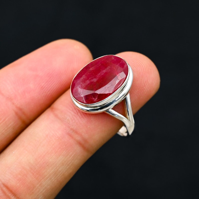 Może przedstawiać: Srebrny pierścionek z dużym, owalnym rubinem. Rubin ma głęboki czerwony kolor i jest osadzony w prostej, eleganckiej srebrnej oprawie. Pierścionek jest prezentowany na czarnym tle.