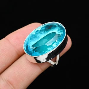 Könnte beinhalten: Ein Ring mit einem großen, oval geschliffenen, hellblauen Edelstein. Der Edelstein hat eine facettierte Oberfläche, die das Licht einfängt und einen funkelnden Effekt erzeugt. Der Ring hat ein silbernes Band.