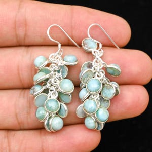 May include: A pair of silver earrings featuring clusters of round, pale blue gemstones. The earrings have a dangling design, with multiple stones connected by delicate silver chains. The gemstones have a smooth, polished appearance, and the earrings are displayed against a neutral background.