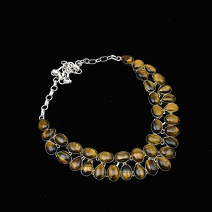 May include: A silver-toned necklace featuring multiple oval and teardrop-shaped tiger's eye gemstones. The stones are a mix of brown and gold hues, set in a double-strand design. The necklace has a chain closure and is set against a black background.