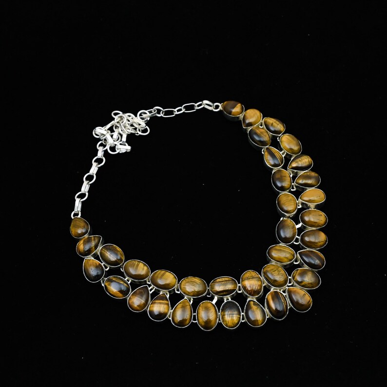 May include: A silver-toned necklace featuring multiple oval and teardrop-shaped tiger's eye gemstones. The stones are a mix of brown and gold hues, set in a double-strand design. The necklace has a chain closure and is set against a black background.