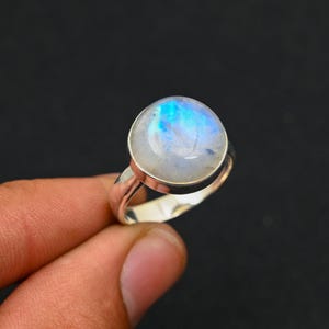 Può includere: Un anello d'argento con una grande pietra di luna rotonda, che mostra sfumature iridescenti blu e bianche. La pietra di luna è incastonata in una lunetta d'argento. La fascia dell'anello è in argento lucido.