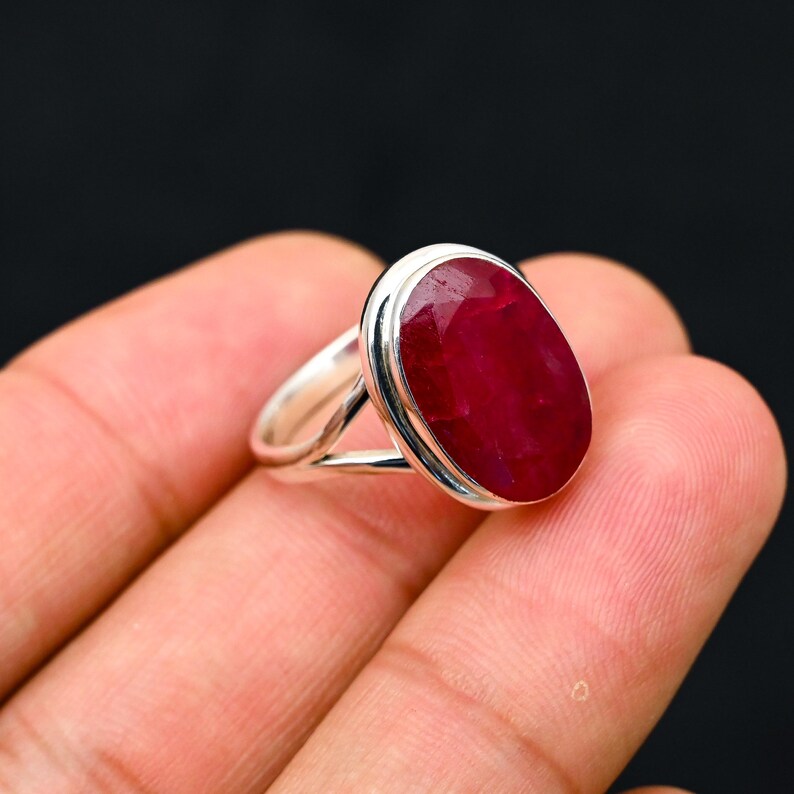 Może przedstawiać: Srebrny pierścionek z dużym, owalnym rubinem. Rubin ma głęboki, bogaty czerwony kolor i jest osadzony w polerowanej srebrnej oprawie. Pierścionek jest prezentowany na ciemnym tle.