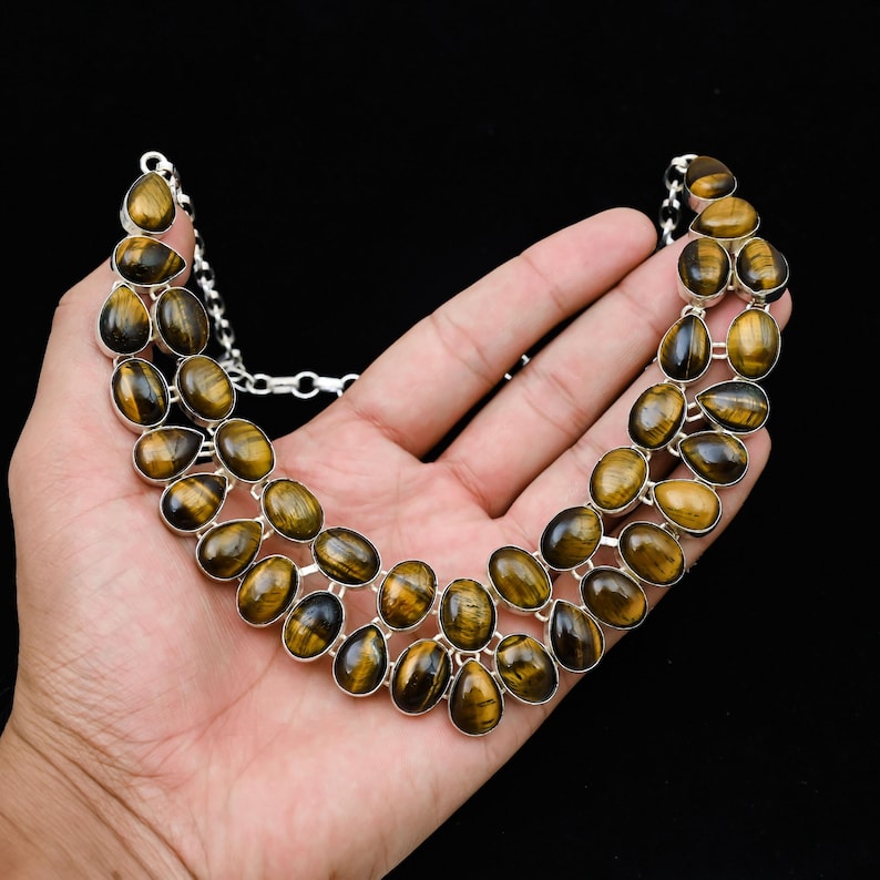 May include: A silver necklace featuring oval-shaped tiger's eye gemstones. The gemstones are a mix of brown and gold, set in a silver metal frame. The necklace is displayed against a black background.