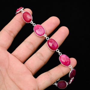 Könnte beinhalten: Ein silbernes Armband mit ovalen, leuchtend roten Edelsteinen. Die Edelsteine sind in einzelnen Silberfassungen gefasst und durch kleine Silberverbinder verbunden. Das Armband wird vor einem dunklen Hintergrund präsentiert.