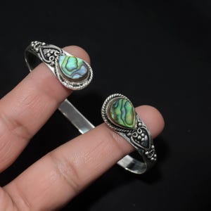 May include: A silver cuff bracelet with two teardrop-shaped abalone shell inlays. The iridescent shells display green, blue, and gold hues. The bracelet features ornate silver detailing around the shell settings and along the band.
