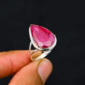 Può includere: Un anello in argento sterling con una grande pietra preziosa di rubino a forma di pera. Il rubino è di un rosso vibrante con una superficie sfaccettata, incastonato in una lunetta d'argento lucida. L'anello ha un design semplice ed elegante.