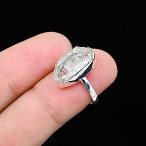Puede incluir: Un anillo de cuarzo transparente con una banda de plata. El cuarzo ovalado tiene inclusiones y está engastado en un bisel. El anillo se muestra sobre un fondo negro.