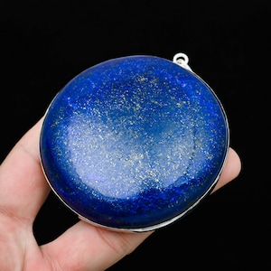 May include: A round, polished lapis lazuli stone with a silver-colored metal rim. The deep blue stone has flecks of gold and a small loop at the top. The background is black.