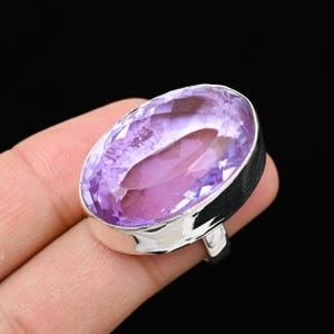 May include: A large, oval-cut amethyst gemstone ring. The amethyst is a light purple color with facets that catch the light. The ring has a silver-colored band.