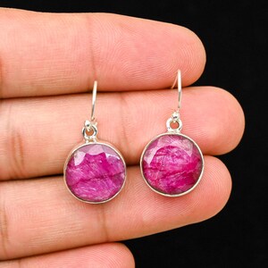 Pode incluir: Um par de brincos pendurados em prata com pedras preciosas redondas, facetadas e magenta. Os brincos têm um design simples com um pequeno laço na parte superior para pendurar. As pedras são fixadas em uma moldura de prata.