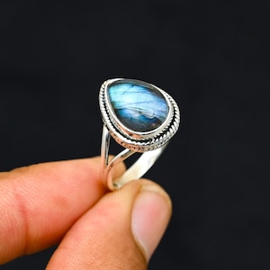 Op de afbeelding: Een zilveren ring met een peervormige labradoriet edelsteen. De steen vertoont iriserende blauwe en groene tinten. De ring heeft een gedetailleerde, touwachtige rand en een gespleten schachtontwerp. Zwarte achtergrond.