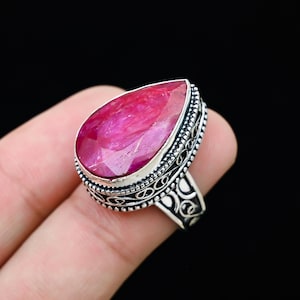 May include: A silver ring featuring a large, pear-shaped, vibrant pink gemstone. The gemstone is the focal point, set in a detailed, ornate silver band with an openwork design. The ring is displayed against a dark background.