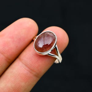 May include: A silver ring featuring a large, oval-shaped, reddish-brown gemstone. The gemstone has a polished, glossy surface. The ring's band is a simple, split-shank design. The ring is set against a dark background.