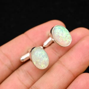 May include: A pair of oval-shaped cufflinks with a silver setting. The cufflinks feature a white opal stone with iridescent green and yellow flecks. The cufflinks are displayed against a dark background.