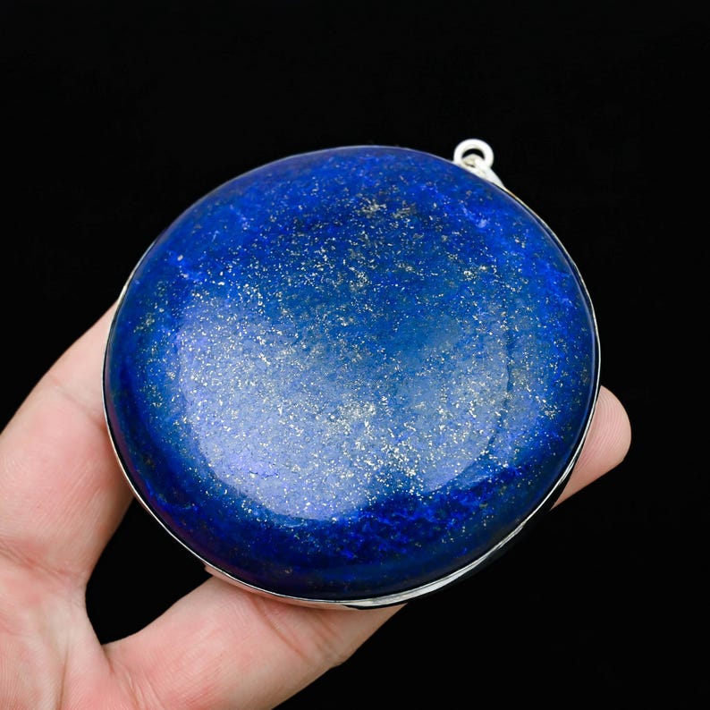 May include: A round, polished lapis lazuli stone with a silver-colored metal rim. The deep blue stone has flecks of gold and a small loop at the top. The background is black.