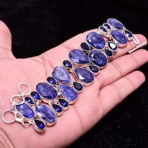 Peut inclure: Un bracelet en argent orné de grandes pierres bleues ovales et en forme de goutte, entrecoupées de petites pierres bleu foncé. Le bracelet est doté d'un fermoir à bascule et est présenté sur une main sur fond noir. Les pierres ont un aspect facetté.