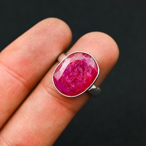 Puede incluir: Un anillo de plata con una gran gema de rubí de corte ovalado. El rubí es de un color rojo vibrante con facetas visibles, engastado en una sencilla banda de plata. El anillo se muestra sobre un fondo oscuro.