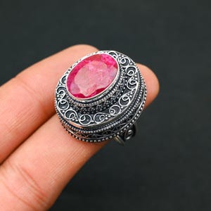 May include: A silver ring featuring a bright pink oval gemstone. The ring has an intricate, detailed design around the gemstone and band. The ring is a piece of jewellery.