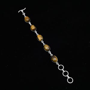 May include: A silver bracelet featuring oval tiger's eye stones. The stones are set in individual silver settings and connected by small silver links. The bracelet has a toggle clasp and three adjustable rings for sizing. The tiger's eye stones have a warm brown color.