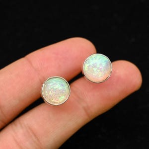 Puede incluir: Un par de pendientes de botón redondos con centros iridiscentes tipo ópalo engastados en metal plateado. Los pendientes tienen un aspecto brillante y multicolor, con tonos de blanco, verde y rosa. Los pendientes se muestran sobre un fondo oscuro.