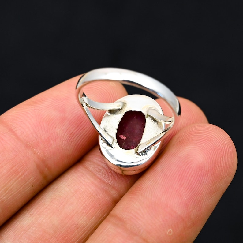 Może przedstawiać: Srebrny pierścionek z owalnym, głęboko czerwonym kamieniem szlachetnym. Pierścionek ma polerowaną srebrną obrączkę, a kamień jest osadzony w oprawie. Pierścionek jest pokazany na ciemnym tle.