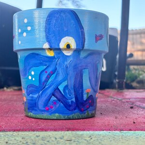Può includere: Vaso di fiori in ceramica blu dipinto a mano con un design di polpo stravagante. Il polpo è dipinto in tonalità di blu con occhi gialli e accenti rossi. Il vaso ha uno sfondo azzurro con bolle bianche e un pesce rosso.