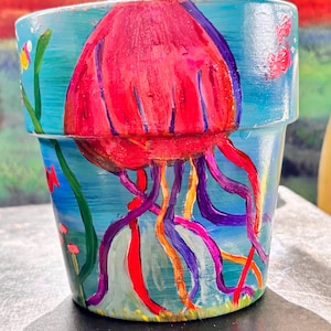 Peut inclure: Pot de fleur peint à la main avec un motif de méduse. Le pot présente une grande méduse rouge avec des tentacules violets, orange et rouges sur un fond bleu. Des algues vertes et du corail rose complètent le thème sous-marin.