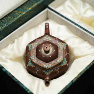 Könnte beinhalten: Eine braune, sechseckige Teekanne mit aufwendigen Blumenmustern in Türkis, Rot und Gold. Die Teekanne wird in einer satin gefütterten Box mit türkisfarbenem und goldfarbenem Zierrat präsentiert, was auf ein hochwertiges Teeservice hindeutet.