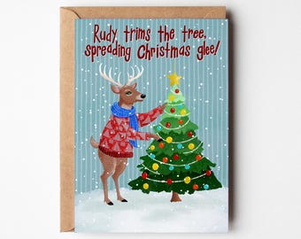 May include: A Christmas card featuring a reindeer wearing a red sweater and blue scarf, decorating a Christmas tree. The card has the text "Rudy trims the tree, spreading Christmas glee!" on a blue background with falling snow.