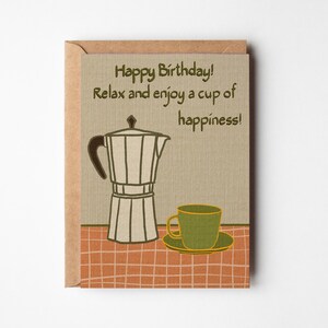 May include: Greeting card with a brown envelope. The card features a coffee maker and a cup of coffee on a checkered background. Text on the card reads: "Happy Birthday! Relax and enjoy a cup of happiness!"