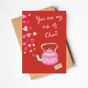 May include: A red card with the text "You are my cup of Chai" in white lettering. A pink teapot with green accents and a floral pattern is illustrated on the card. Pink hearts are illustrated around the teapot.
