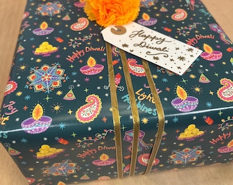 May include: A square gift box wrapped in dark teal paper featuring a colorful pattern of diya lamps, paisley shapes, and the text Happy Diwali. The box is decorated with three gold ribbons, an orange carnation, and a white gift tag reading Happy Diwali.