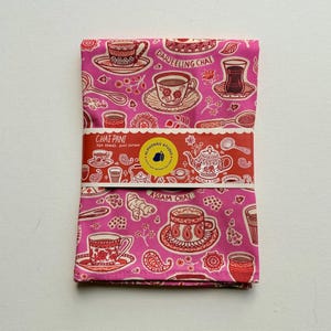May include: A folded pink tea towel with a repeating pattern of teacups, teapots, and spoons in red and white. The towel is wrapped with a red band that reads "CHAI-PANT" and features a logo.