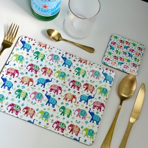 May include: A set of colourful elephant-themed placemats and coasters. The placemats and coasters feature a white background with a repeating pattern of colourful elephants and flowers. The placemats are rectangular and the coasters are square.