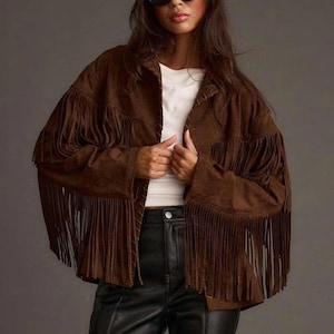May include: A brown suede jacket with long fringe detailing. The jacket has a collared neckline and is worn over a white top and black trousers. The model is wearing black sunglasses.