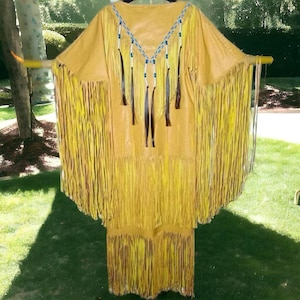 May include: A mustard-yellow leather garment with long fringe detailing. The top features a V-neck with blue and white bead accents and black feather-like embellishments. The garment is displayed outdoors.