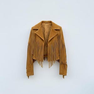Puede incluir: Chaqueta de ante marrón claro con flecos. La chaqueta tiene un cuello clásico, mangas largas y un largo recortado. Los flecos están a lo largo de las mangas y la parte delantera. La chaqueta se muestra sobre un fondo blanco.