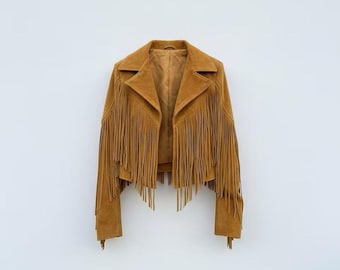 May include: A tan suede jacket with fringe detailing. The jacket has a classic collar, long sleeves, and a cropped length. The fringe is along the sleeves and the front. The jacket is displayed against a white background.