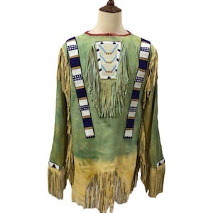Könnte beinhalten: Ein Vintage-Hemd im indianischen Stil mit langen Ärmeln in Grün- und Beigetönen. Das Hemd hat dekorative Perlenarbeiten in Weiß, Blau und Gelb mit langen Fransen an den Ärmeln und am Saum. Das Hemd wird auf einer Schaufensterpuppe präsentiert.