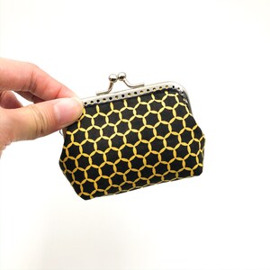 Puede incluir: Una pequeña cartera de monedas negra con un patrón geométrico dorado. La cartera tiene un cierre de metal.