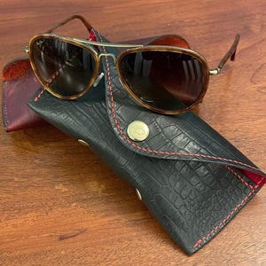 May include: Aviator sunglasses with brown frames and dark lenses sit on a black, textured leather case with red stitching. The case has a gold button closure and a gold emblem. A burgundy leather case is visible.