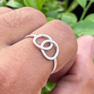 May include: A silver ring featuring a double-circle design. The ring is held up against a backdrop of green foliage. The ring's minimalist design is simple and elegant.