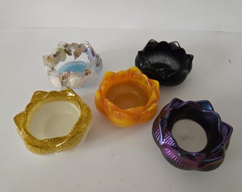 May include: Five decorative candle holders in different colours and designs. One is black, one is gold glitter, one is orange, one is iridescent purple, and one is clear with seashells. Each holder has a unique shape.
