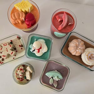 May include: A selection of decorative candles in glass containers. The candles are shaped like pumpkins, leaves, gingerbread men, and a snowman. The colours are orange, red, green, and white, creating a festive and seasonal theme.