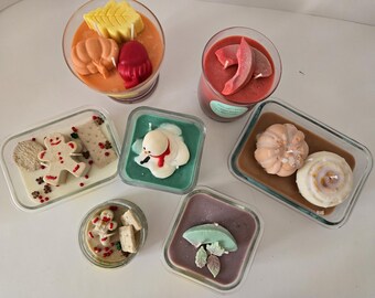 May include: A selection of decorative candles in glass containers. The candles are shaped like pumpkins, leaves, gingerbread men, and a snowman. The colours are orange, red, green, and white, creating a festive and seasonal theme.