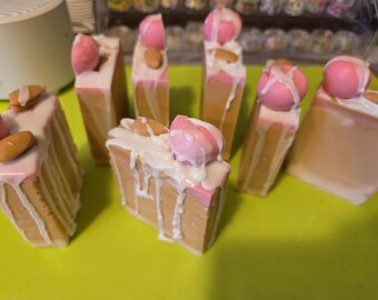 May include: A selection of cake-slice-shaped soaps. The soaps have a light brown base, a pink layer, and white icing drizzled over the top. Some are decorated with pink spheres and almond slivers.
